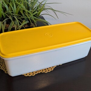 May include: A rectangular white plastic storage container with a bright yellow snap-on lid. The lid features a centered circular embossed logo. The container has smooth, straight sides and rounded corners, providing a clean and functional design.