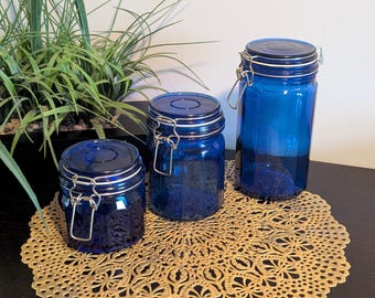 May include: Three cobalt blue glass jars with silver metal lids and clasps. The jars are sitting on a gold lace doily.