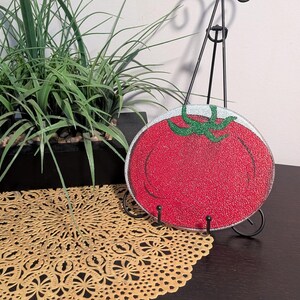 May include: A round, clear glass cutting board with a red tomato design. The tomato has a green glitter stem and a white outline. The cutting board is displayed on a black metal stand, with a decorative gold lace doily in the foreground.