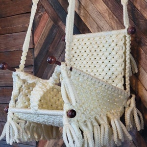 May include: Cream-colored macrame hanging basket with a woven design and fringe detailing. The basket features wooden dowels and a knotted rope for hanging. A decorative storage solution for a bohemian or vintage-inspired space.