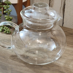 May include: A clear glass teapot with a lid. The teapot has a round body and a handle. The teapot is sitting on a wooden surface.