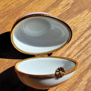 May include: An open, egg-shaped trinket box with a pink lid and white interior. The box is trimmed with gold and features a small, gold flower accent. The box is on a wooden surface.