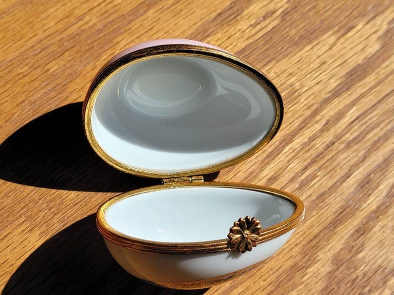 May include: An open, egg-shaped trinket box with a pink lid and white interior. The box is trimmed with gold and features a small, gold flower accent. The box is on a wooden surface.