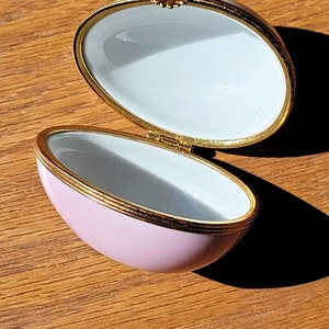May include: Open, oval-shaped trinket box with a light pink exterior and a white interior. The box has gold trim and a small floral accent on the hinge. The box is on a wooden surface.