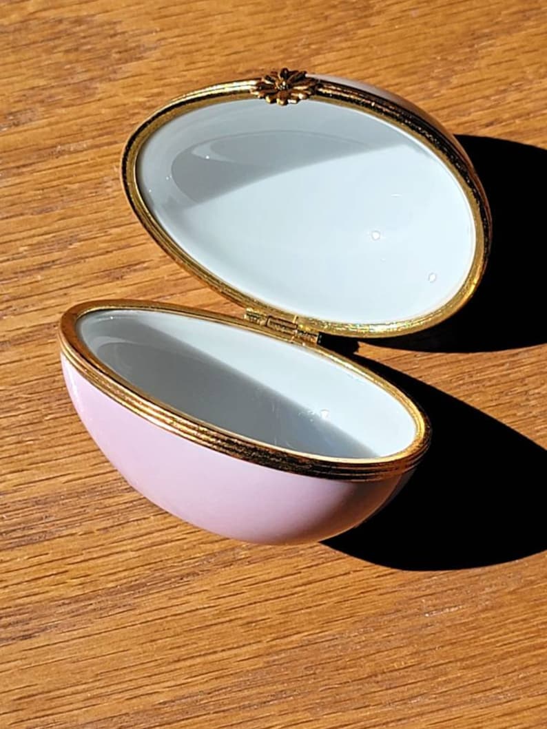 May include: Open, oval-shaped trinket box with a light pink exterior and a white interior. The box has gold trim and a small floral accent on the hinge. The box is on a wooden surface.