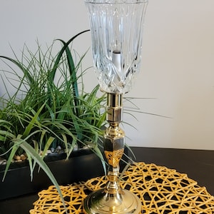 May include: A gold candlestick with a clear glass shade. The candlestick is on a woven placemat.