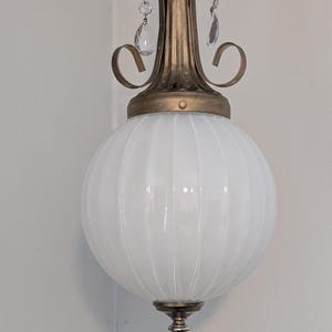 May include: A vintage white globe pendant light with vertical ribbed detailing. The light fixture has a brass-colored top with decorative scrollwork and hanging crystal accents. The bottom features a decorative silver-colored finial.