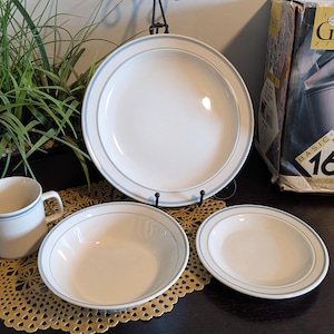 May include: A white ceramic dinnerware set with a light blue border. The set includes a large plate, a bowl, a smaller plate, and a mug. A box with the text "Gibson" is visible in the background.