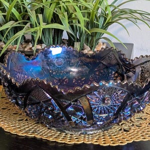 May include: An iridescent, dark blue glass bowl with a ruffled edge. The bowl features intricate geometric and floral patterns. The bowl sits on a gold-colored, lacy placemat, with greenery in the background.