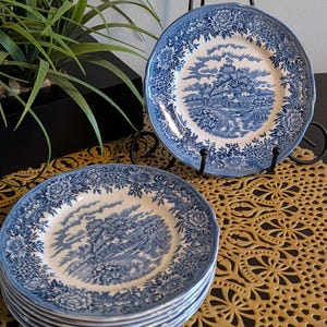 May include: A stack of blue and white porcelain plates with a floral border and a landscape scene in the center. One plate is displayed on a black metal stand. The plates are on a gold-colored, patterned placemat.