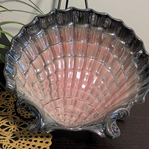 May include: A decorative, iridescent seashell-shaped bowl with a silver-toned rim and base. The interior has a pinkish hue, and the bowl is supported by two ornate, curved feet. The bowl is likely made of glass or a similar material.
