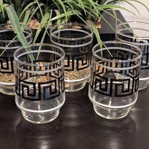 May include: Set of five clear drinking glasses with a black geometric design and white trim. The glasses are cylindrical with a slightly flared base. The design is a repeating pattern of black squares and lines. The glasses are arranged on a dark surface.