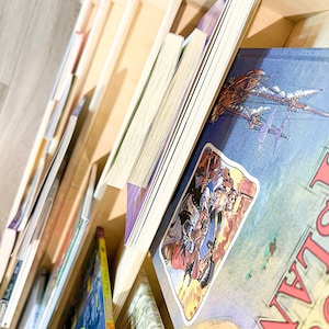 May include: A wooden bookshelf filled with various books. The books have colorful covers and different sizes. Some books are standing upright, while others are lying down. The bookshelf is made of light-colored wood.