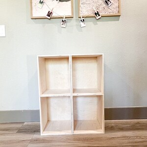 May include: A light wood shelf unit with four open cubbies. The shelf unit is standing on a light wood floor.