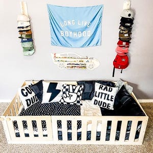 May include: A wooden toddler bed with a black and white checkered bedding set. The bed has a wooden railing around the perimeter. There are three pillows with the words "Cool Dude", "Rad Little Dude", and a peace sign on them. There is a blue flag with the words "Long Live Boyhood" hanging on the wall behind the bed.
