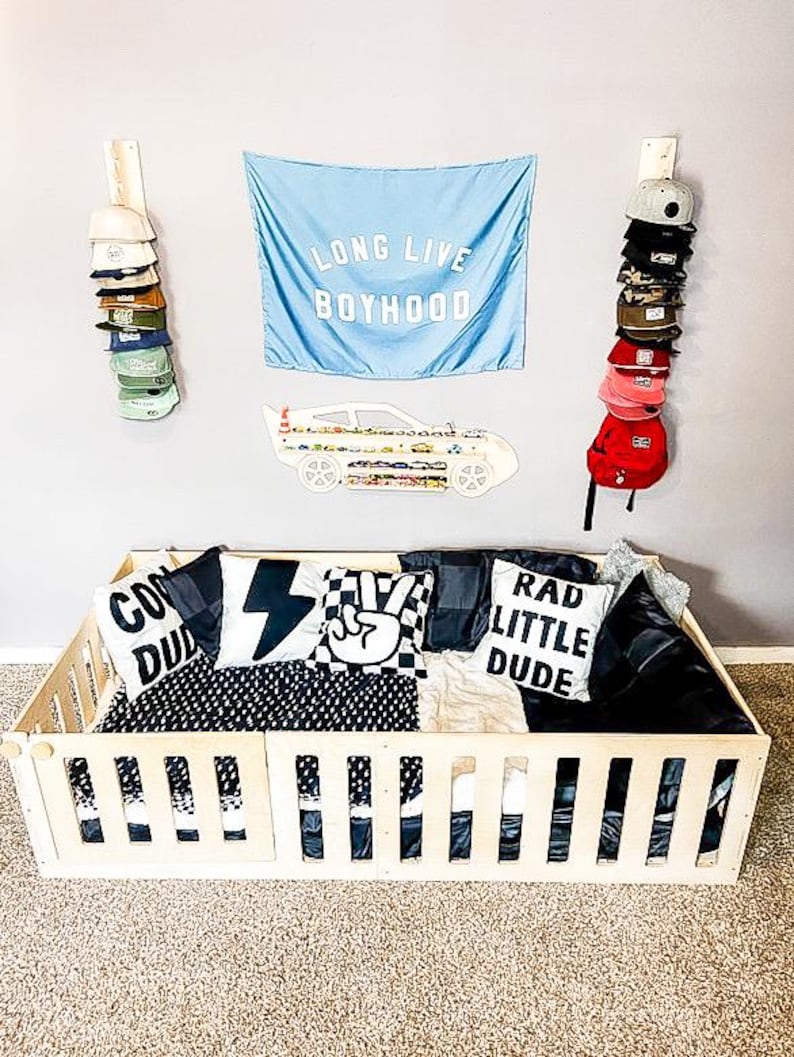 May include: A wooden toddler bed with a black and white checkered bedding set. The bed has a wooden railing around the perimeter. There are three pillows with the words "Cool Dude", "Rad Little Dude", and a peace sign on them. There is a blue flag with the words "Long Live Boyhood" hanging on the wall behind the bed.