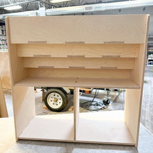 May include: A light-colored, unfinished wooden display shelf with multiple shelves and compartments. The shelf has a rectangular shape with rounded edges and is constructed from plywood. The design includes several horizontal shelves and a divided lower section.