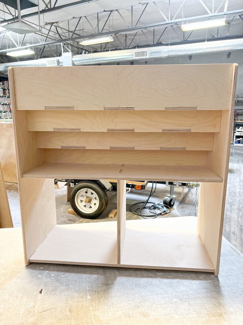 May include: A light-colored, unfinished wooden display shelf with multiple shelves and compartments. The shelf has a rectangular shape with rounded edges and is constructed from plywood. The design includes several horizontal shelves and a divided lower section.
