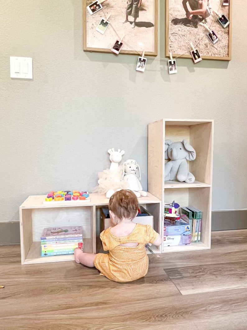 May include: A wooden storage unit with three shelves, a toddler sitting in front of it, and a variety of toys and books on the shelves. The unit is made of light wood and has a simple design.
