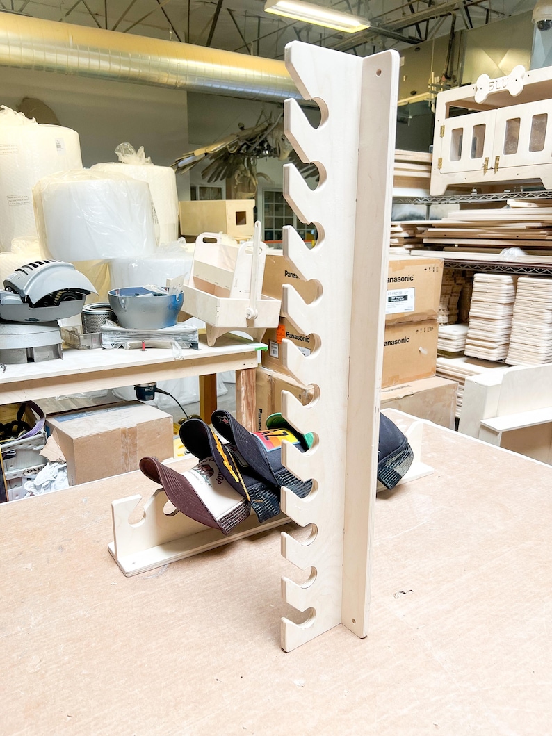 May include: A wooden hat rack with 10 slots for storing baseball caps. The rack is made of light-colored wood and has a simple design. There are several baseball caps hanging on the rack.