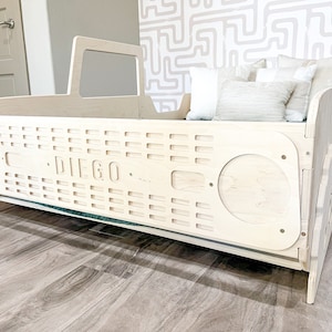 Puede incluir: Una cama de madera para niños pequeños con forma de camión y acabado blanco. La cama tiene un recorte de una parrilla con la palabra "DIEGO" en ella. La cama está sobre un suelo de madera marrón claro.
