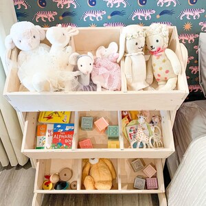 May include: A white wooden toy shelf with three shelves. The top shelf has stuffed toys, the middle shelf has books and blocks, and the bottom shelf has toys and blocks.