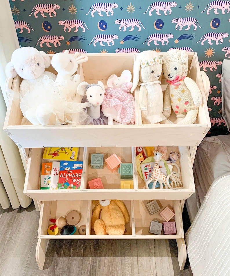 May include: A white wooden toy shelf with three shelves. The top shelf has stuffed toys, the middle shelf has books and blocks, and the bottom shelf has toys and blocks.