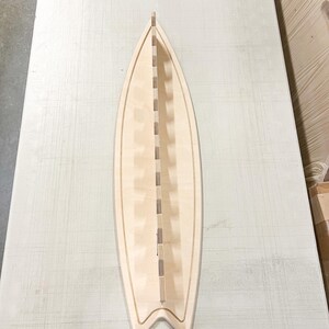 May include: A wooden surfboard rack with slots for storing multiple surfboards. The rack is made of light-colored wood and has a surfboard shape.