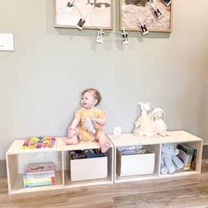 May include: A set of three wooden storage bins with open fronts, each containing various toys and books. A young child sits on the bins, holding a stuffed animal. The bins are arranged in a row on a wooden floor.