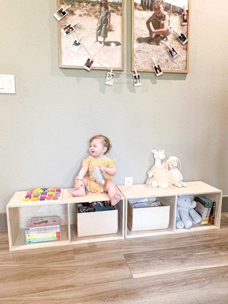 May include: A set of three wooden storage bins with open fronts, each containing various toys and books. A young child sits on the bins, holding a stuffed animal. The bins are arranged in a row on a wooden floor.