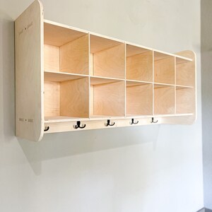 May include: A light-colored wooden storage unit with multiple cubby holes and a row of black hooks below. The unit is mounted on a light gray wall, offering a functional and stylish storage solution for any space.