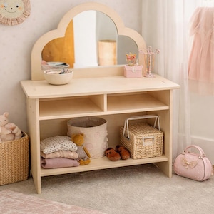May include: A light wood children's vanity with a cloud-shaped mirror. The vanity has shelves with baskets, folded clothes, and a teddy bear. A pink purse and rug are in the foreground. A woven sun-shaped wall hanging is on the wall.