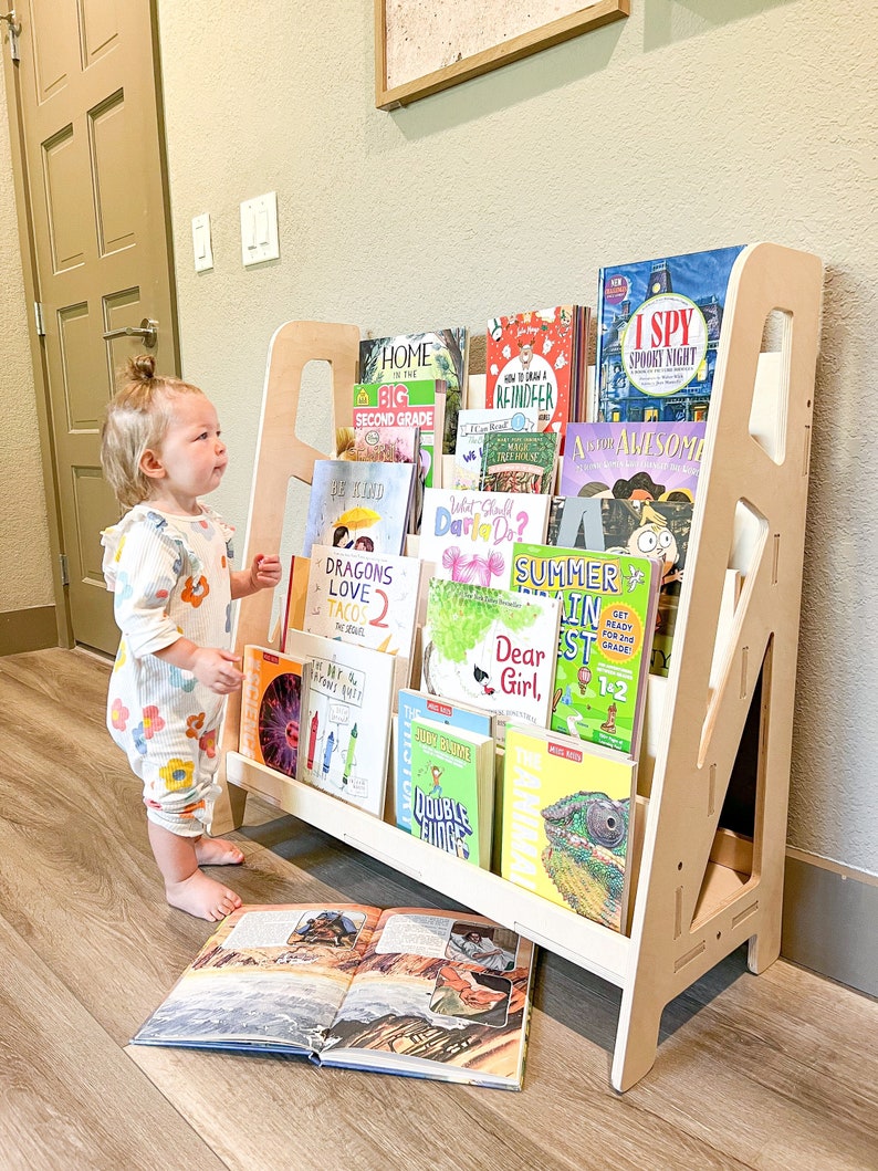 May include: A light-colored wooden bookshelf filled with children's books. The bookshelf has a unique A-frame design. A young child in a floral jumpsuit stands near the bookshelf, looking at an open book on the floor.