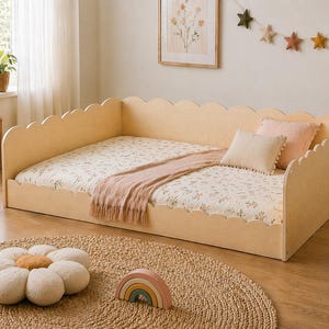 May include: A light wood-toned bed frame with a scalloped headboard and footboard. The bed is made up with a floral patterned sheet, a pink throw blanket, and several pillows. A large daisy-shaped floor cushion and a wooden rainbow are in the foreground.