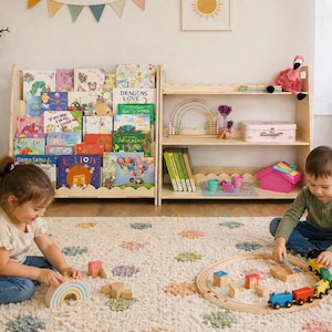 May include: Two children playing with toys in a bright room. A wooden bookshelf is filled with colorful children's books. A wooden train set and a rainbow-shaped toy are on the floor. A pink flamingo decoration sits on a shelf.