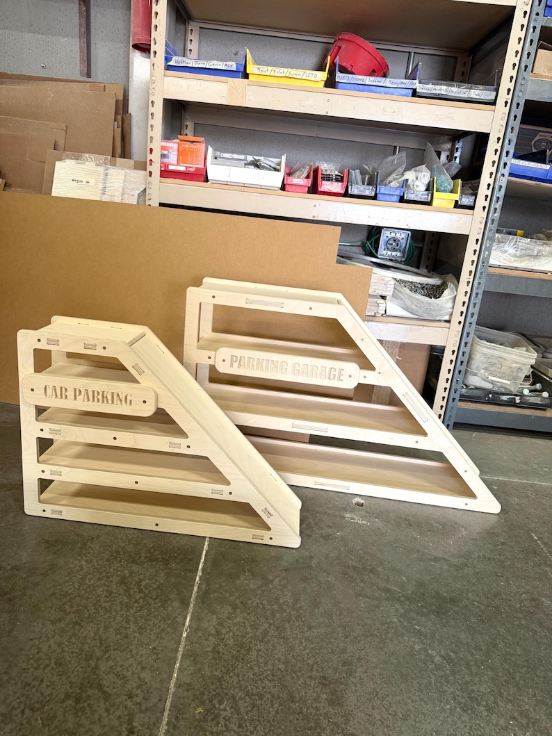 May include: Two wooden car parking garages. One is labeled "CAR PARKING" and the other is labeled "PARKING GARAGE". Both have a natural wood finish and a triangular design with multiple shelves for toy cars.