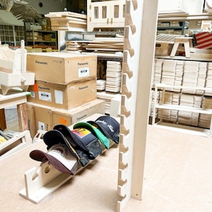 May include: A wooden hat rack with six slots for storing hats. The rack is made of light-colored wood and has a natural finish. Five baseball caps are displayed on the rack, including a green cap with the text "Cowboy Style" in white.