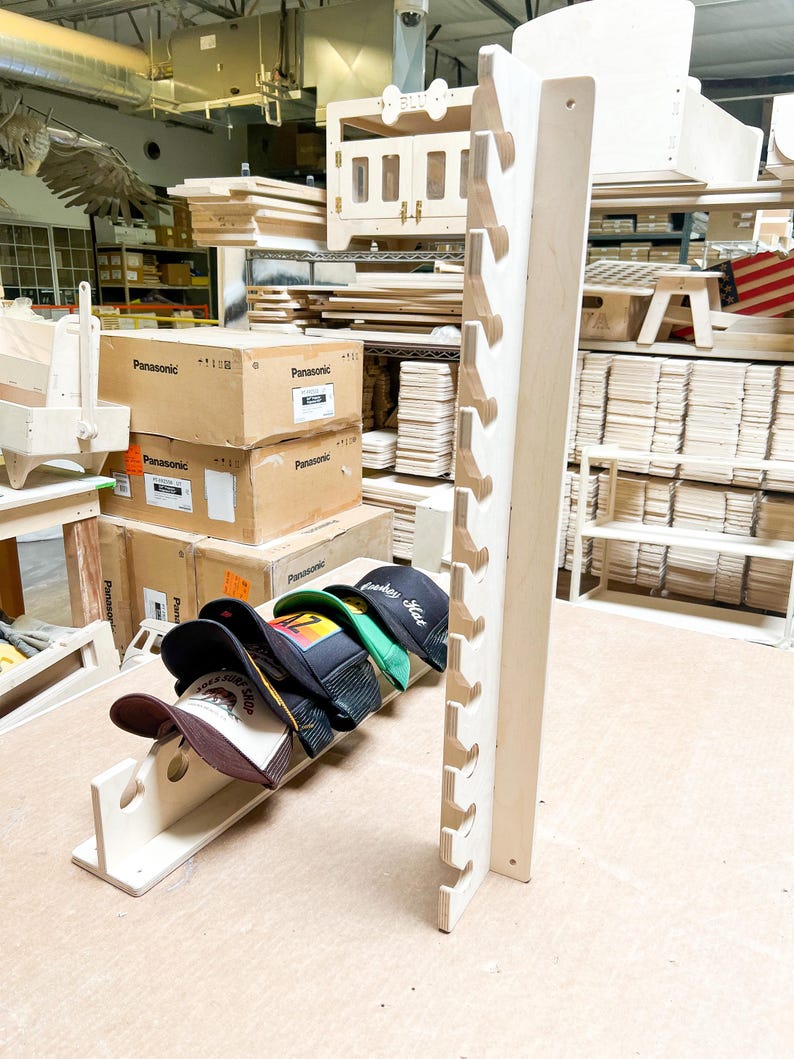 May include: A wooden hat rack with six slots for storing hats. The rack is made of light-colored wood and has a natural finish. Five baseball caps are displayed on the rack, including a green cap with the text "Cowboy Style" in white.