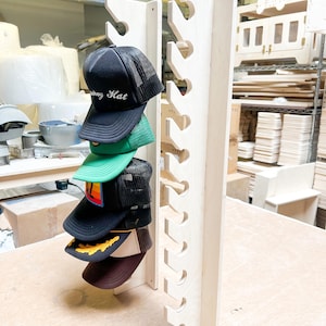 May include: A wooden hat rack with 10 slots for storing baseball caps. The rack is made of light wood and has a natural finish. Five baseball caps are displayed on the rack, including one with the text "Subway Hat" embroidered on the front.