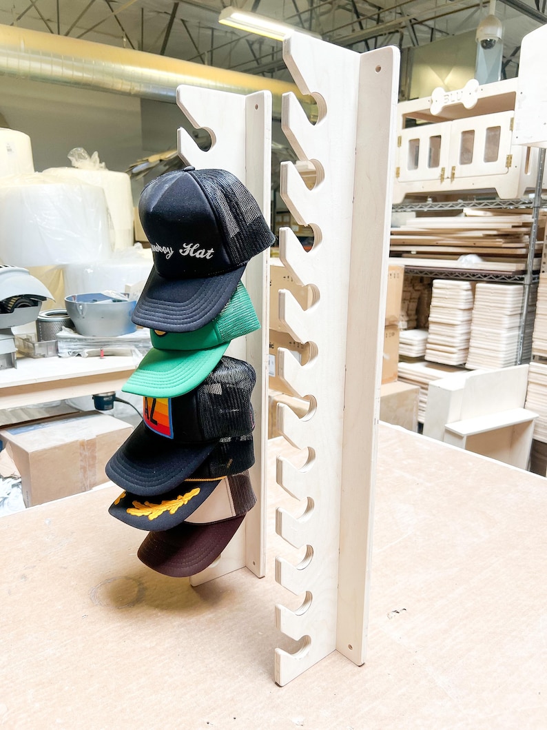 May include: A wooden hat rack with 10 slots for storing baseball caps. The rack is made of light wood and has a natural finish. Five baseball caps are displayed on the rack, including one with the text "Subway Hat" embroidered on the front.