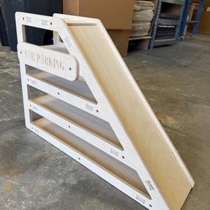 May include: A wooden car parking ramp with a natural wood slide and white frame. The words "CAR PARKING" are engraved on the top. The ramp has three parking levels and is designed for toy cars.