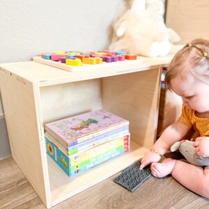 May include: A wooden storage cube with a shelf and a stack of colourful children's books. A child is sitting on the floor playing with a grey textured toy.