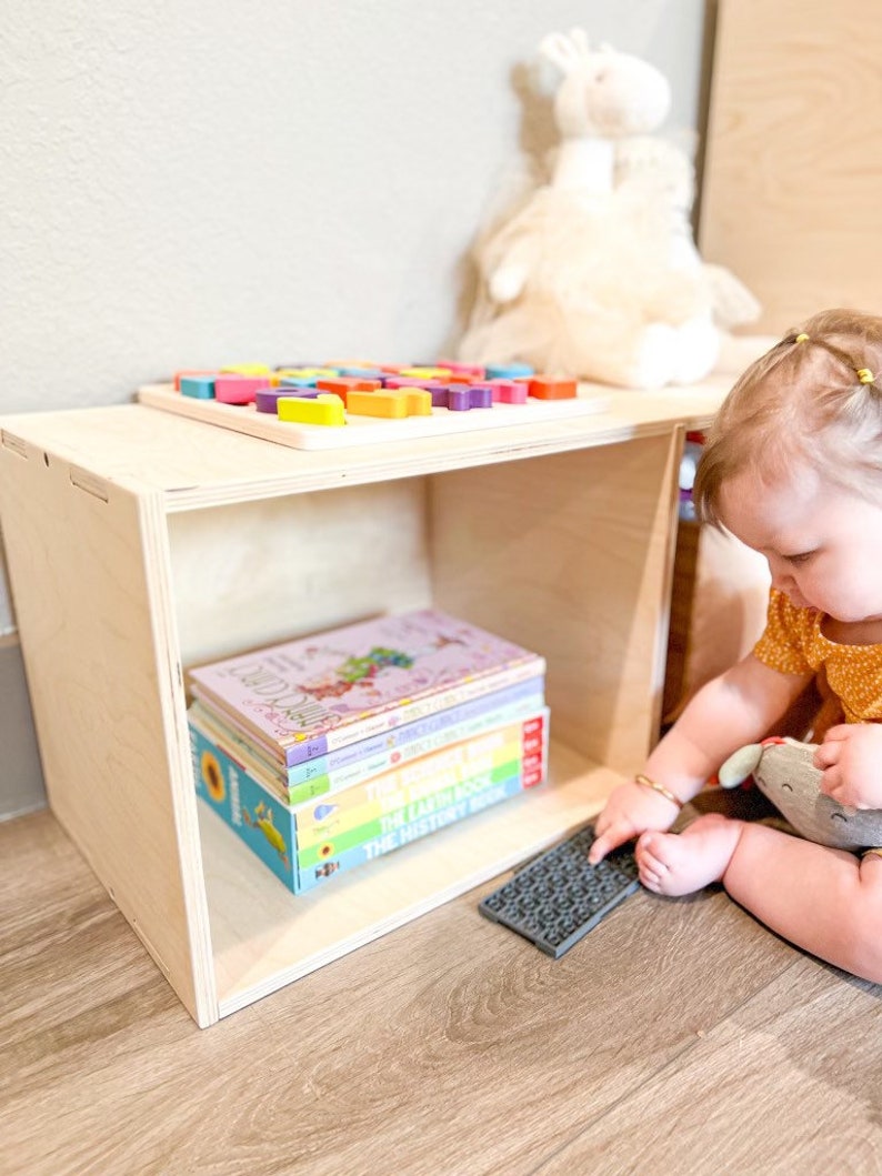 May include: A wooden storage cube with a shelf and a stack of colourful children's books. A child is sitting on the floor playing with a grey textured toy.