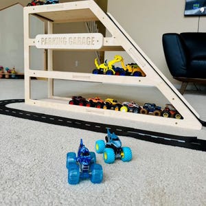 May include: A wooden parking garage with multiple levels, holding various toy trucks and cars. The sign on the garage reads "PARKING GARAGE". A black road is in front of the garage, and two blue monster trucks are in the foreground.