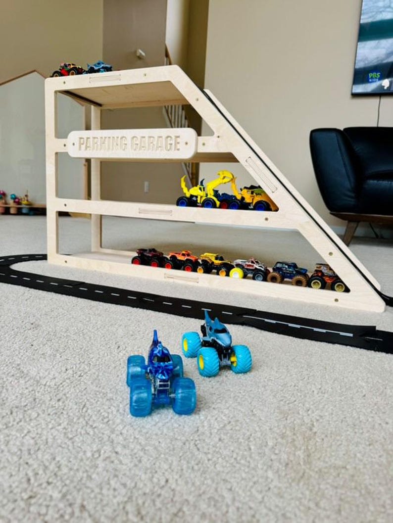 May include: A wooden parking garage with multiple levels, holding various toy trucks and cars. The sign on the garage reads "PARKING GARAGE". A black road is in front of the garage, and two blue monster trucks are in the foreground.