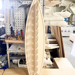 May include: A light wood surfboard-shaped wall rack with multiple slots for hanging items. The rack is made of plywood and has a natural wood finish.