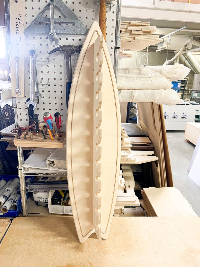 May include: A light wood surfboard-shaped wall rack with multiple slots for hanging items. The rack is made of plywood and has a natural wood finish.