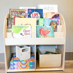 May include: A light-colored wooden bookshelf filled with children's books. The bookshelf has multiple shelves and compartments, with books of various sizes and colors. There are also two small wooden boxes and a colorful box with geometric patterns.