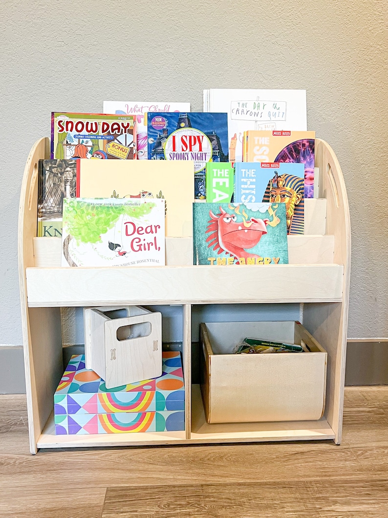 May include: A light-colored wooden bookshelf filled with children's books. The bookshelf has multiple shelves and compartments, with books of various sizes and colors. There are also two small wooden boxes and a colorful box with geometric patterns.