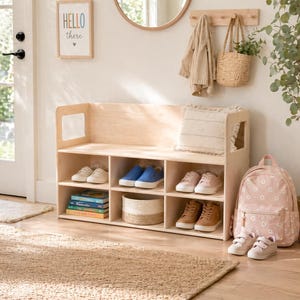 May include: A light wood shoe bench with cubby storage, holding various pairs of shoes, books, and a woven basket. A pink floral backpack and a pair of sneakers sit nearby. A sign that says "Hello there" hangs on the wall.